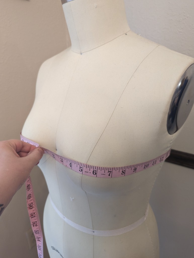 A hand holds a pink tape measure around a dress form at the fullest part of the bust.