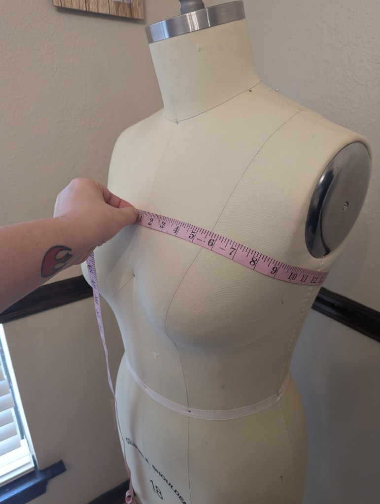 A hand holds a pink measuring tape at the high bust measurement on an armless dress form.