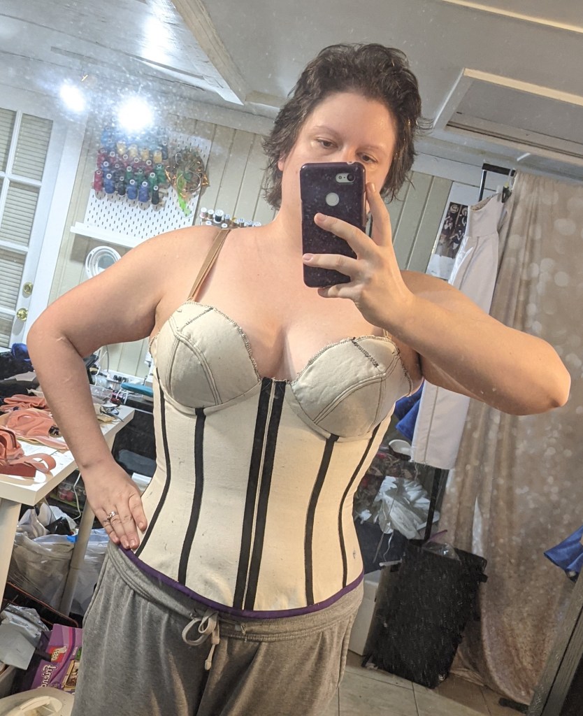 Mirror selfie of TGS wearing a beige cupped corset and grey sweatpants.